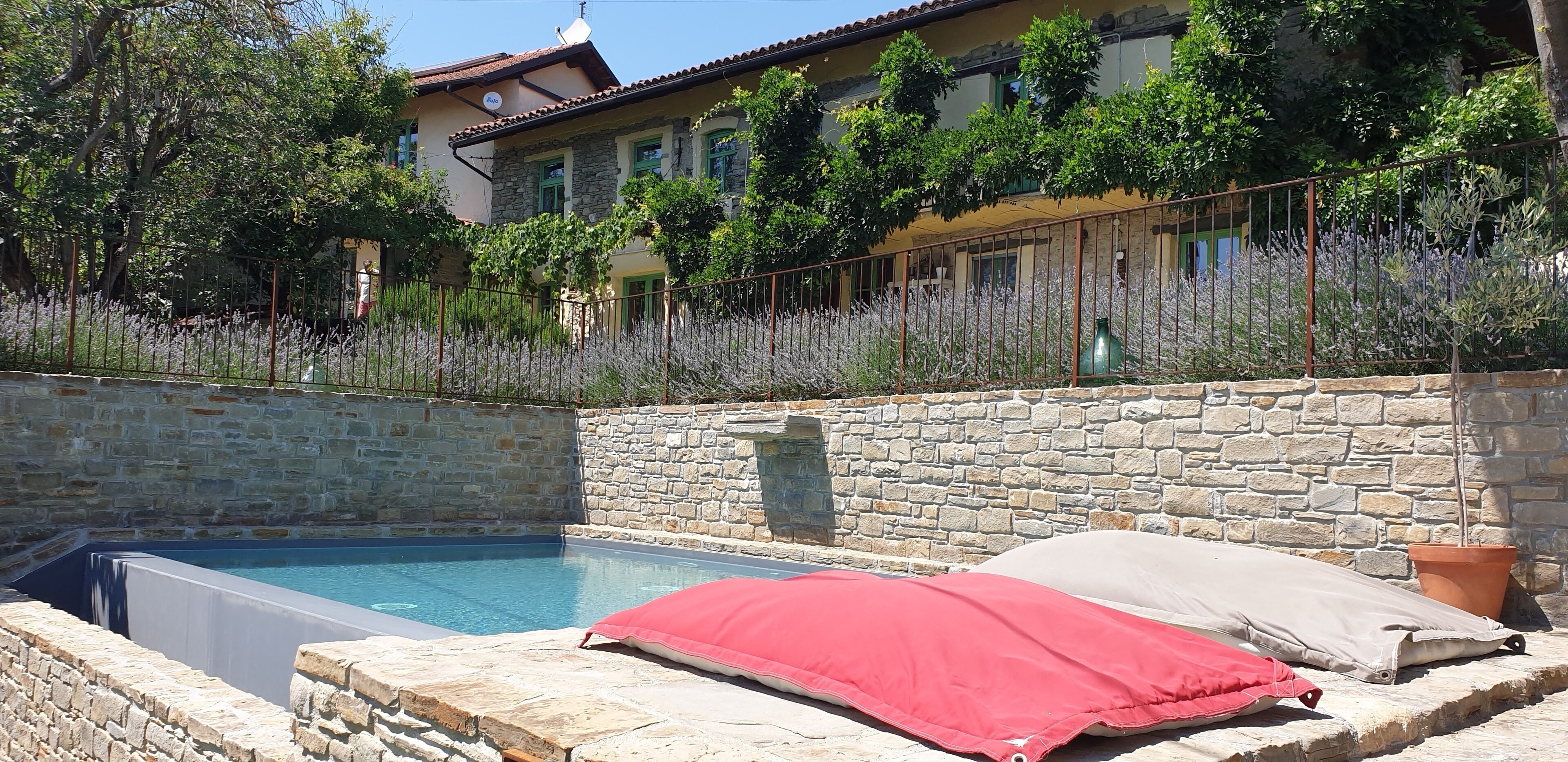 A stone-walled pool with a red and beige cushion, surrounded by a garden with lavender plants and a stone building in the background.