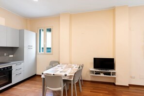 Eine moderne Küche mit weißen Schränken, einem Esstisch mit Stühlen, einem Fernseher auf einem Schrank und einem Fenster mit Jalousien.