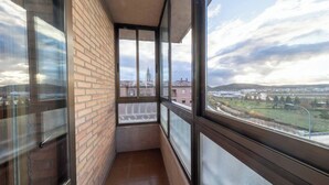 A narrow balcony with a brick wall and large windows offering a view of buildings and a distant landscape.