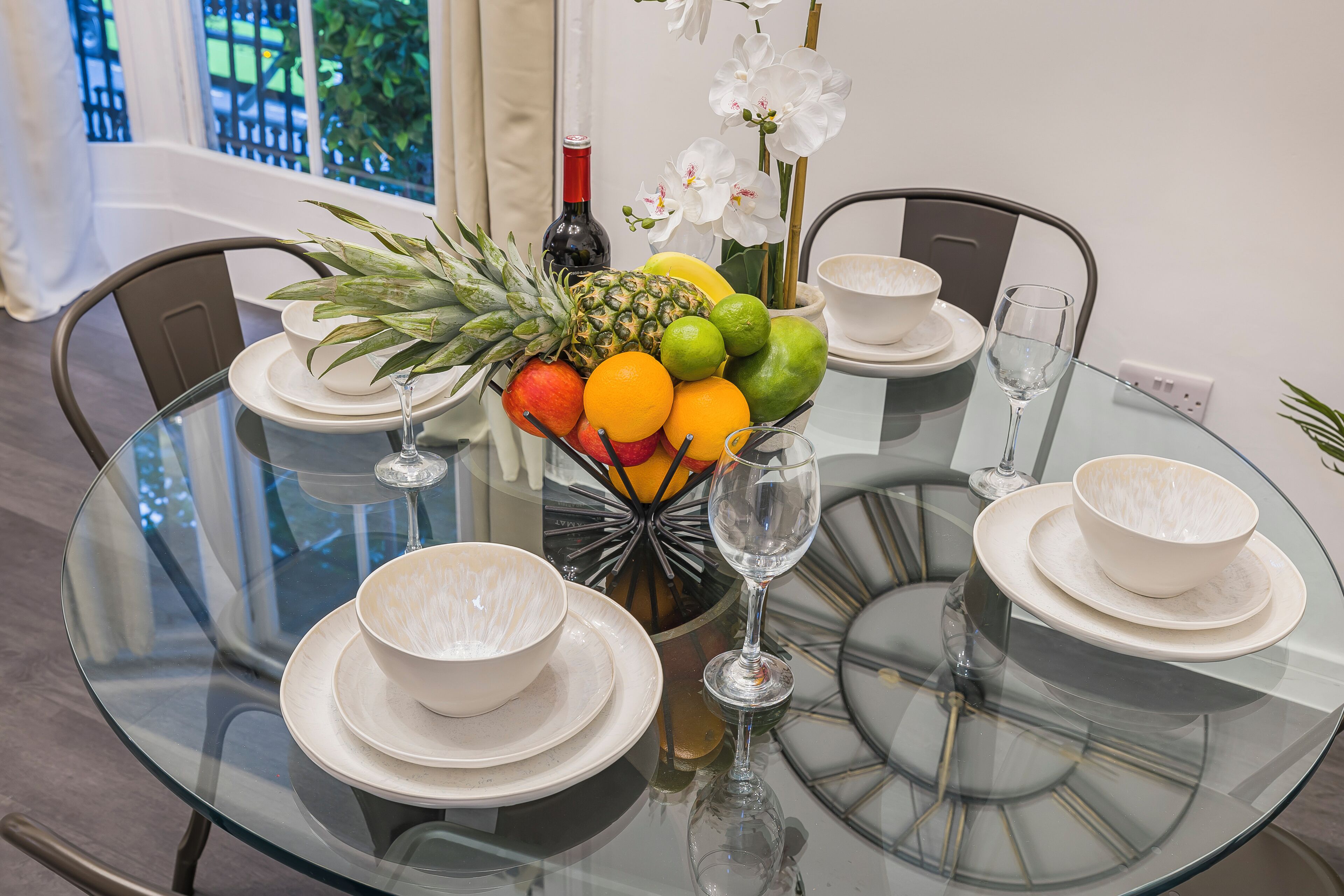 A round glass table with white plates, bowls, and a glass of wine. A fruit bowl with pineapple, oranges, and limes is on the table.