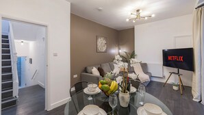 A modern living room with a dining table set for two, a sofa, a TV displaying 'NETFLIX', and a staircase leading to another room. - Station Side Sanctuary - 2BR Central Brighton Stay (Brighton and Hove)