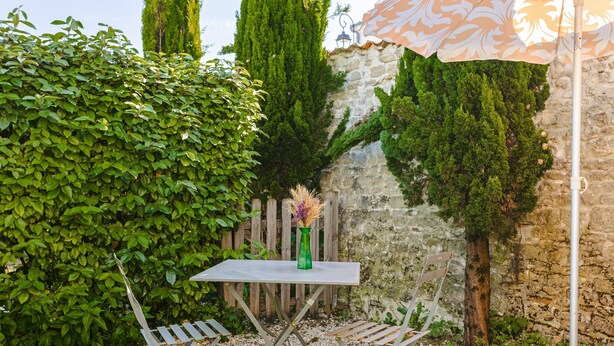 A small outdoor seating area with a table, two chairs, a decorative vase, and a patterned umbrella. - Charming Gîte "Les Jardins d'Harmonie" for 2 in Bourgneuf – Charente-Maritime (Bourgneuf)