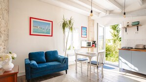 A modern living room with a blue sofa, white dining table, and chairs, a kitchen area with a stove and shelves, and a large window with a view of greenery.