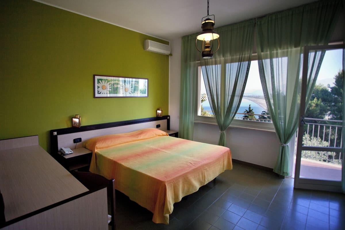 Un dormitorio con cama, mesita de noche, lámpara, un cuadro en la pared y ventana con vistas.