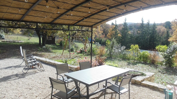 Un patio exterior con mesa y sillas, rodeado de un área de grava y un jardín con árboles y arbustos.