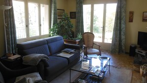 Un salón con un sofá, una silla, una mesa de centro, un televisor y un gran ventanal con cortinas.