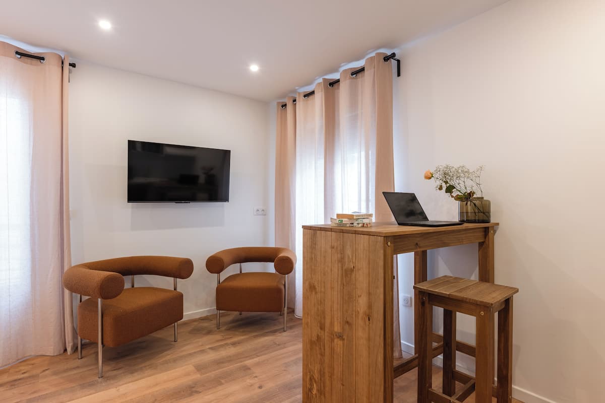 A modern living room with a flat-screen TV mounted on the wall, two brown armchairs, a wooden coffee table with a laptop and decorative items, and light-colored curtains.