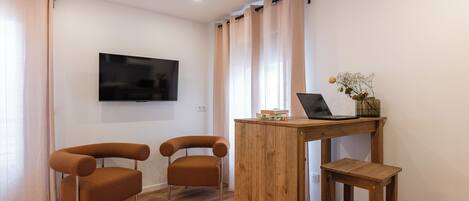 A modern living room with a flat-screen TV mounted on the wall, two brown armchairs, a wooden coffee table with a laptop and decorative items, and light-colored curtains.