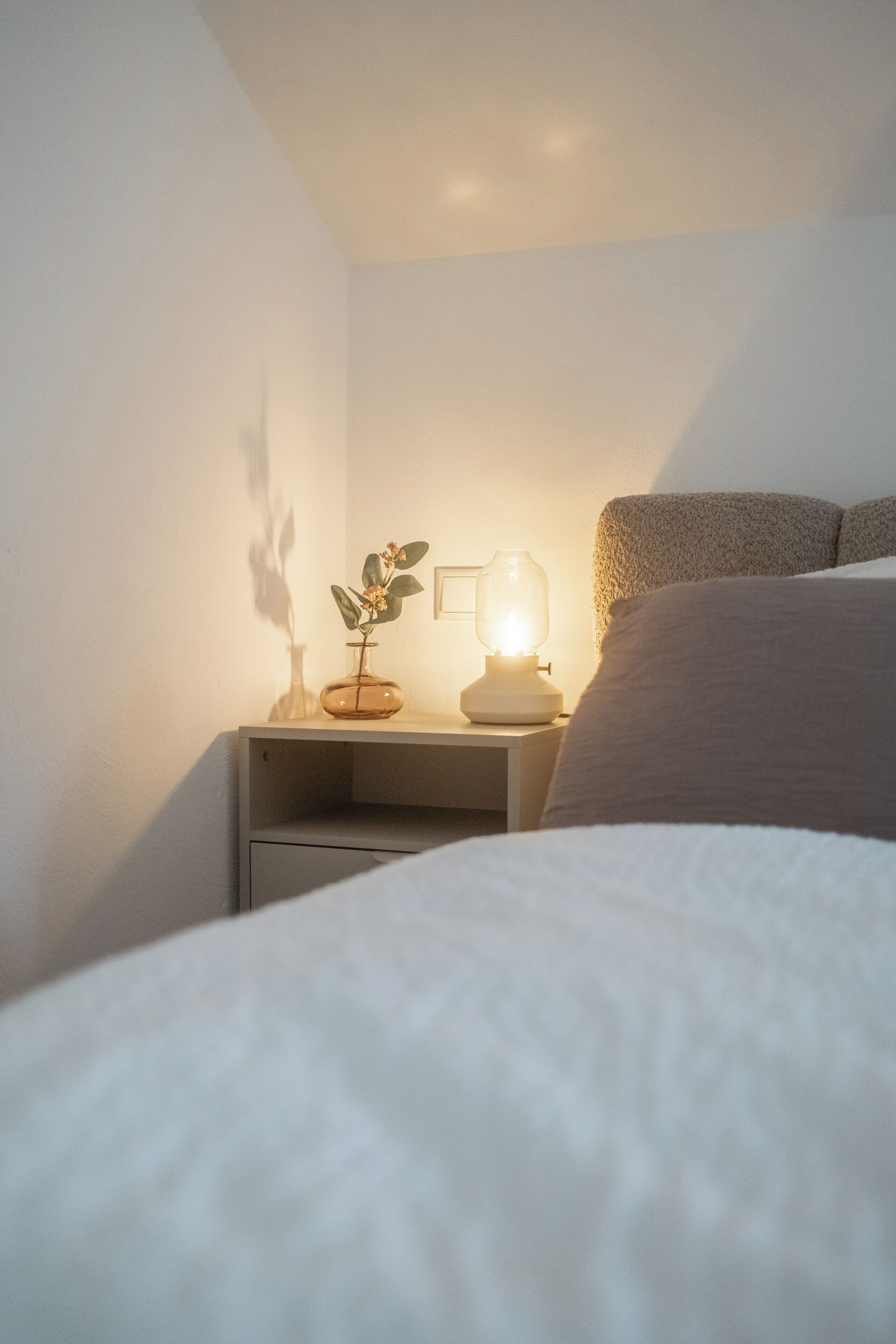 A bedroom with a bed, a bedside table, a lamp, and a plant.