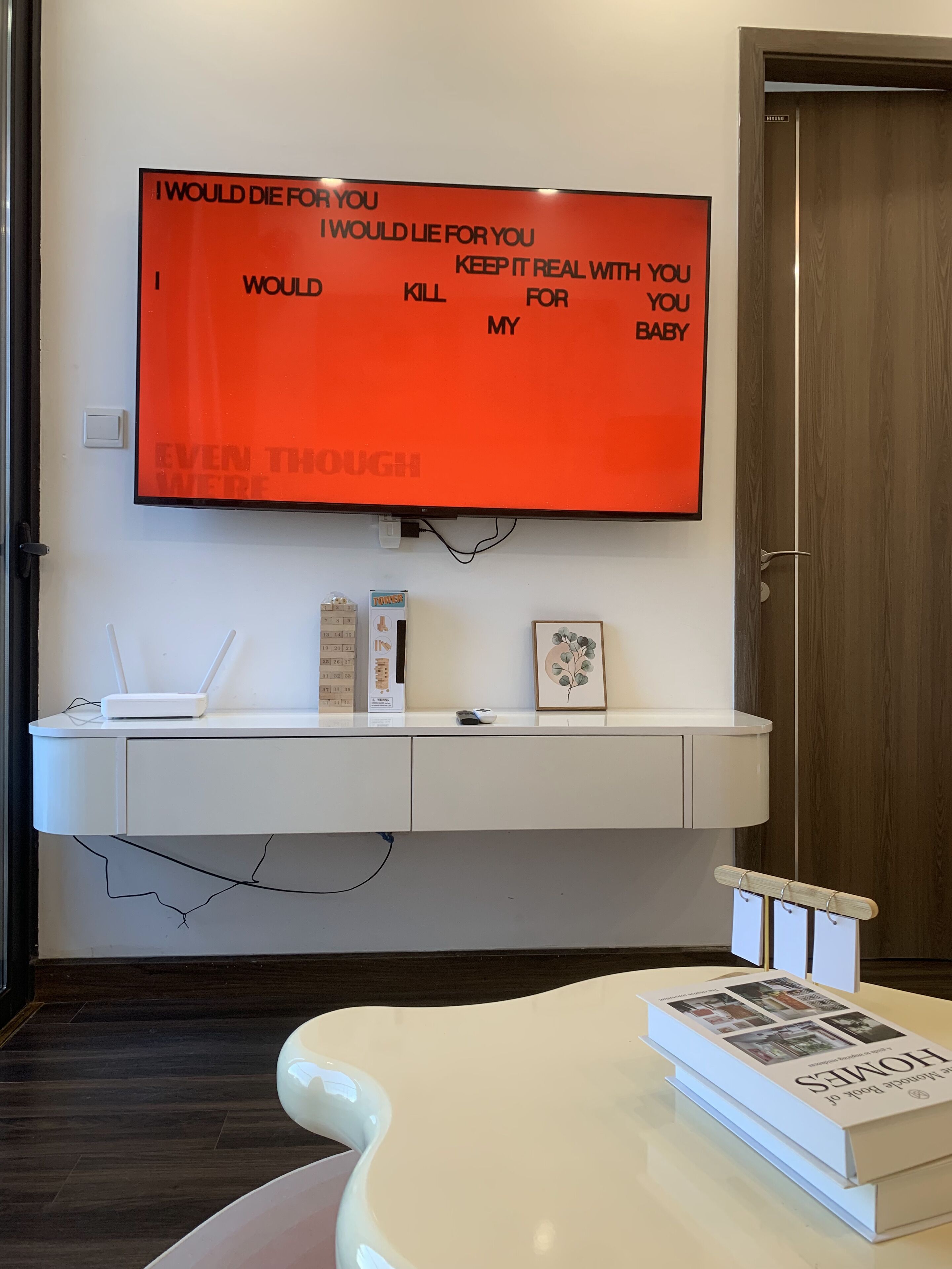A modern room with a mounted TV displaying a motivational quote, a white console table with a router and a framed picture, and a white coffee table with a book titled "HOMES".