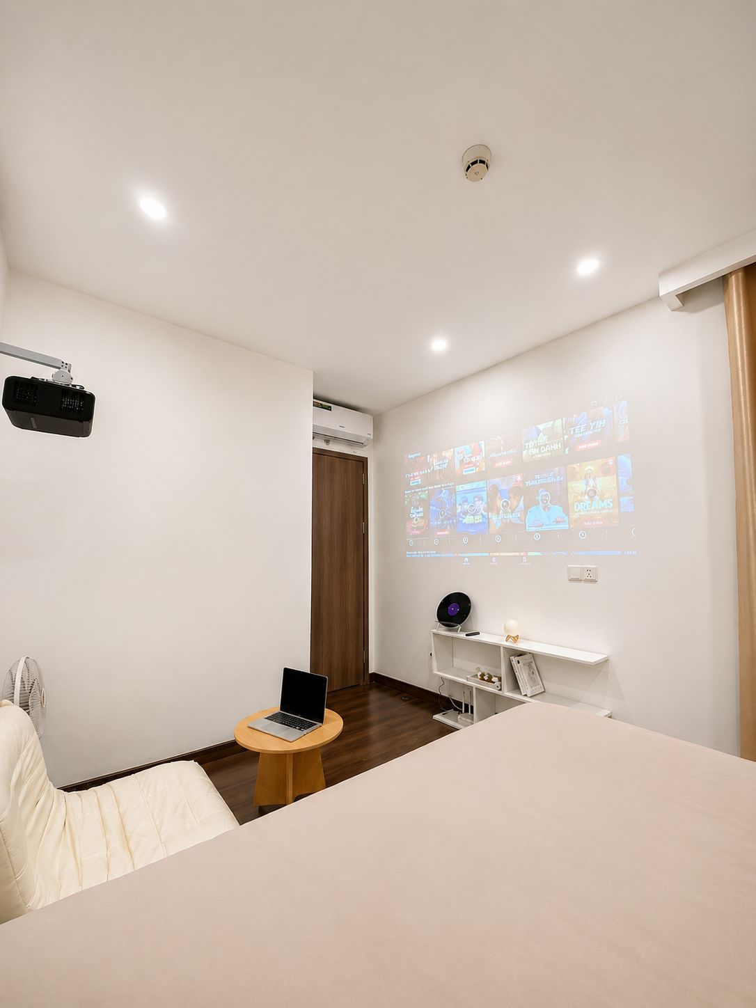A hotel room with a bed, a desk with a laptop, a projector screen displaying content, and a ceiling-mounted air conditioning unit.