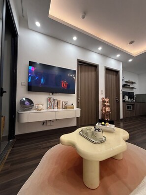 Uma sala de estar moderna com televisão de tela plana na parede, uma mesa de centro bege com um tabuleiro de xícaras e uma área de cozinha ao fundo.