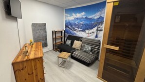 Ein Zimmer mit einem Holzschrank, einer schwarzen Couch mit gemusterten Kissen, einem kleinen Holztisch und einer großflächigen Wandmalerei einer verschneiten Berglandschaft.