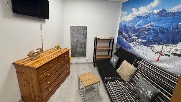 Ein modernes Zimmer mit einem hölzernen Schminktisch, einer schwarzen Couch mit gestreiften Kissen, einem kleinen Tisch, einem an der Wand befestigten Fernseher und einer großen Wandmalerei mit Berglandschaft.