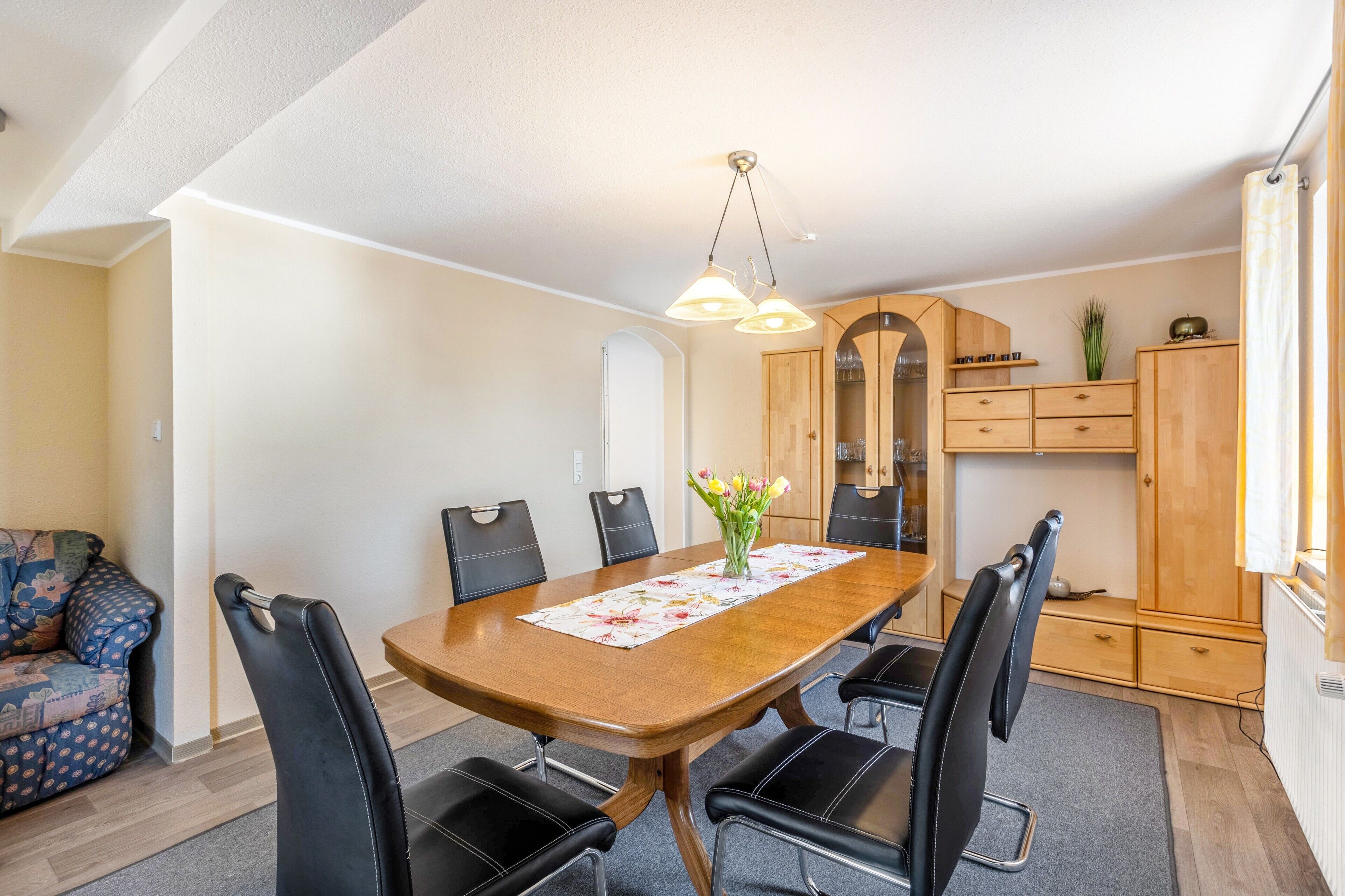 A dining area with a wooden table, black chairs, a vase with flowers, and a ceiling light fixture.