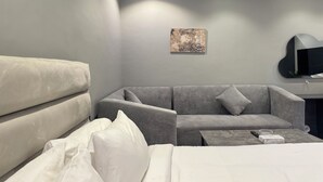 Une chambre d’hôtel moderne avec un canapé gris, un lit avec du linge de lit blanc, une petite table d’appoint et un téléviseur fixé au mur.