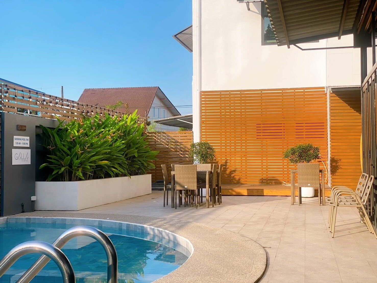 Una piscina con cerco de madera y sillas, rodeada de plantas en macetas y un edificio con pared de listones.