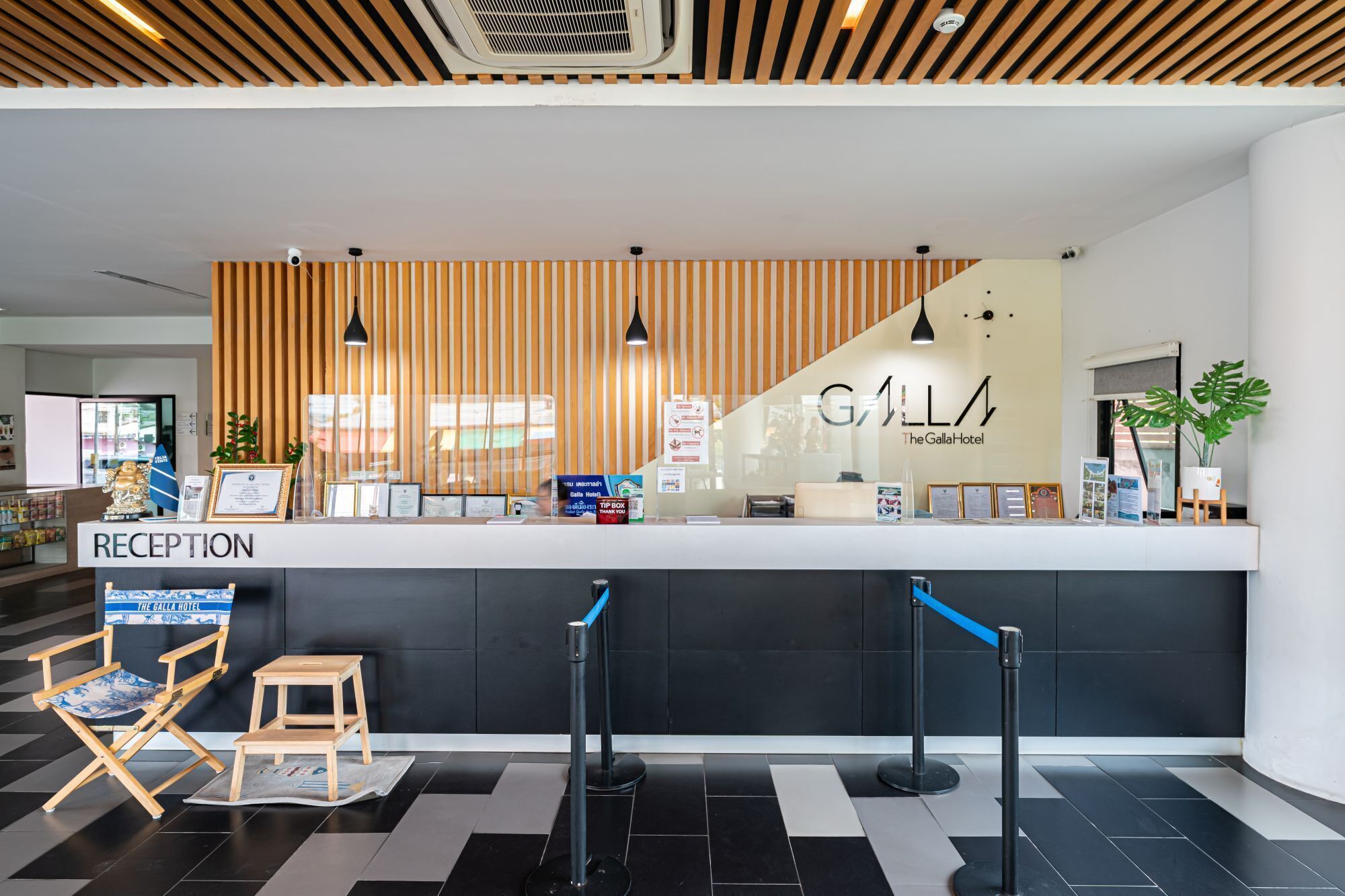 A reception area with a wooden slat wall, a reception desk, and a 'GALLA Hotel' sign. There are two wooden chairs and a small table, a blue rope barrier, and a potted plant.