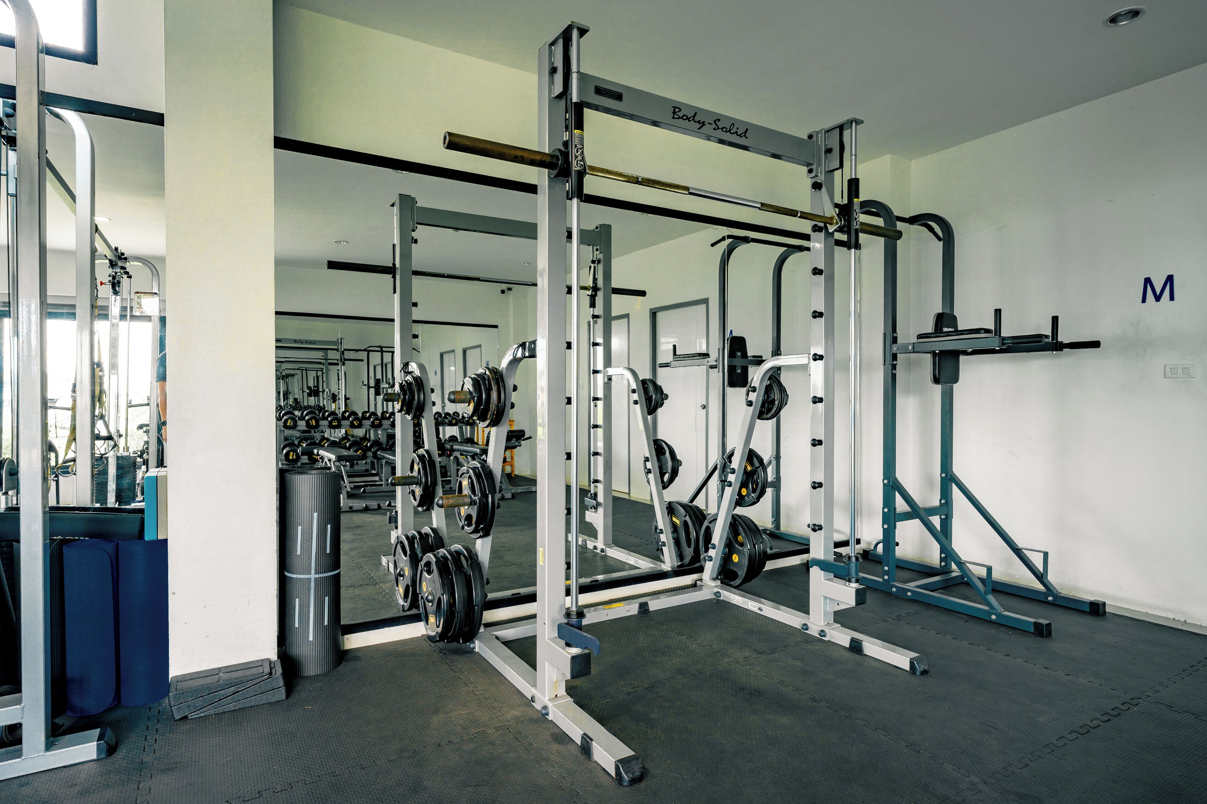 A gym with various weightlifting equipment, mirrors, and a wall with a letter 'M'.