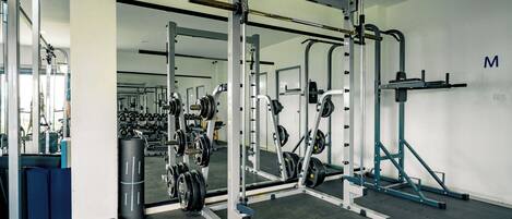 A gym with various weightlifting equipment, mirrors, and a wall with a letter 'M'.