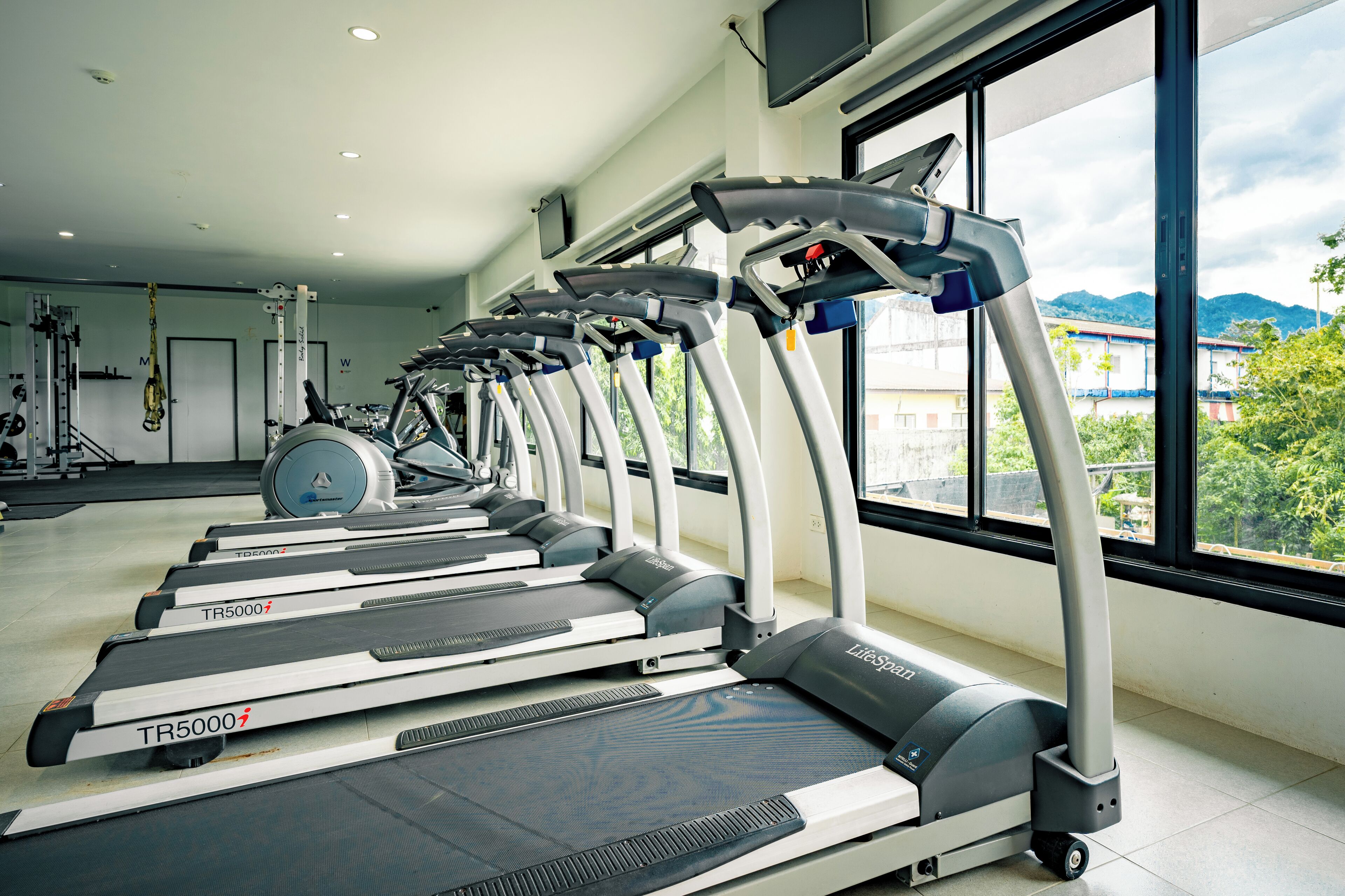 A row of treadmills in a gym with large windows and a view of buildings and mountains.