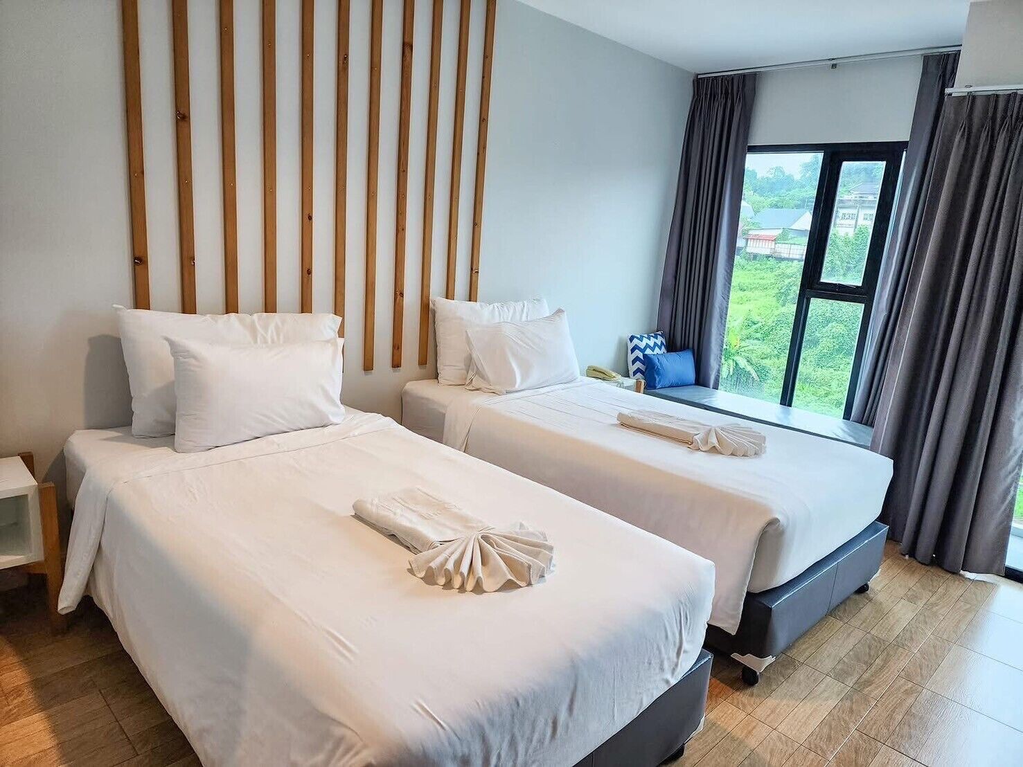 Une chambre d’hôtel avec deux lits, une tête de lit en lattes de bois, une grande fenêtre avec des rideaux et une vue sur la verdure.