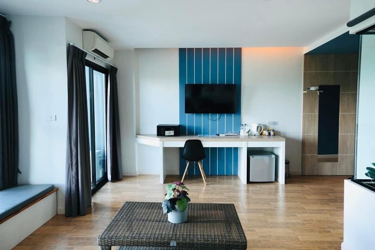 A modern hotel room with a kitchenette, a flat-screen TV mounted on the wall, a small dining table with chairs, and a potted plant on a wicker ottoman.