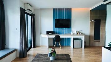 A modern hotel room with a kitchenette, a flat-screen TV mounted on the wall, a small dining table with chairs, and a potted plant on a wicker ottoman.
