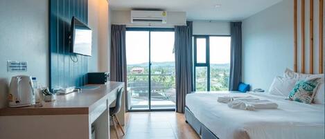 A modern hotel room with a large bed, a desk with a chair, a flat-screen TV, and a view of the city through the windows.