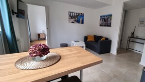A modern living room with a wooden dining table, a black sofa with orange cushions, and a small white side table. There are two framed pictures on the wall, a potted plant, and a trash bin in the corner. - Bright apartment with terrace in the heart of Morlaix (Morlaix)