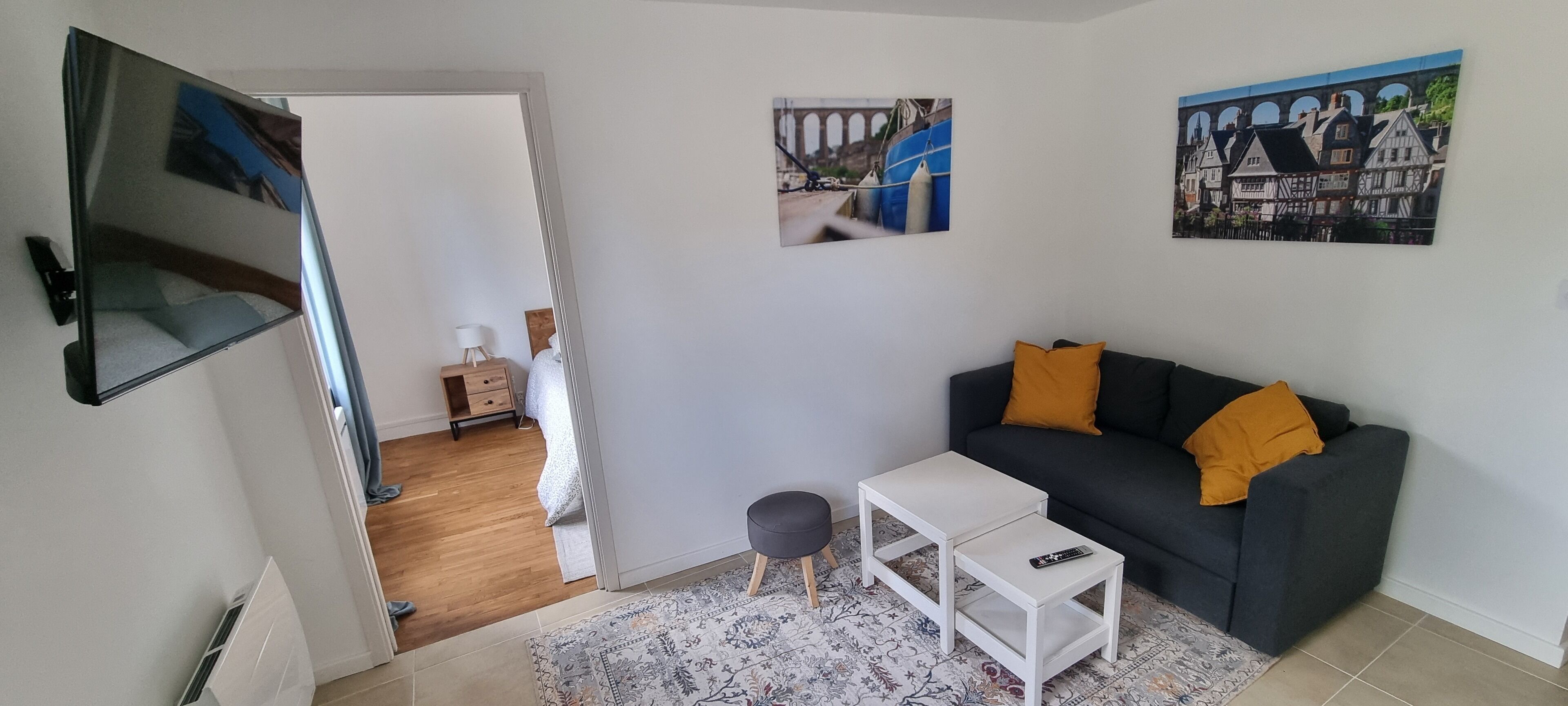 Un salon avec un canapé gris foncé, une table d’appoint blanche et un tapis à motifs. Deux tableaux sont accrochés au mur et représentent un pont et des maisons.