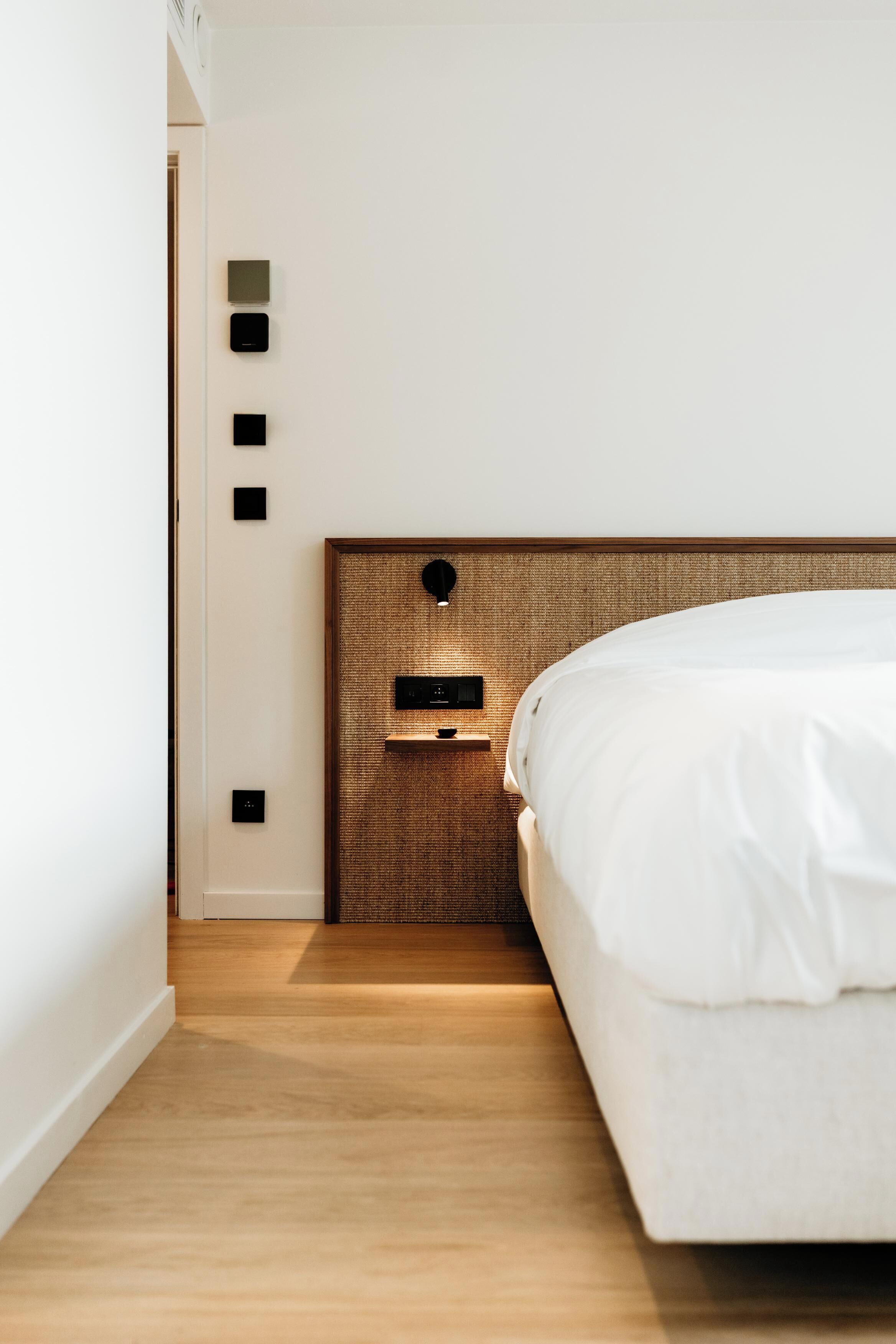 Un dormitorio moderno con cabecera de madera, cama blanca y mesita de noche con iluminación integrada.