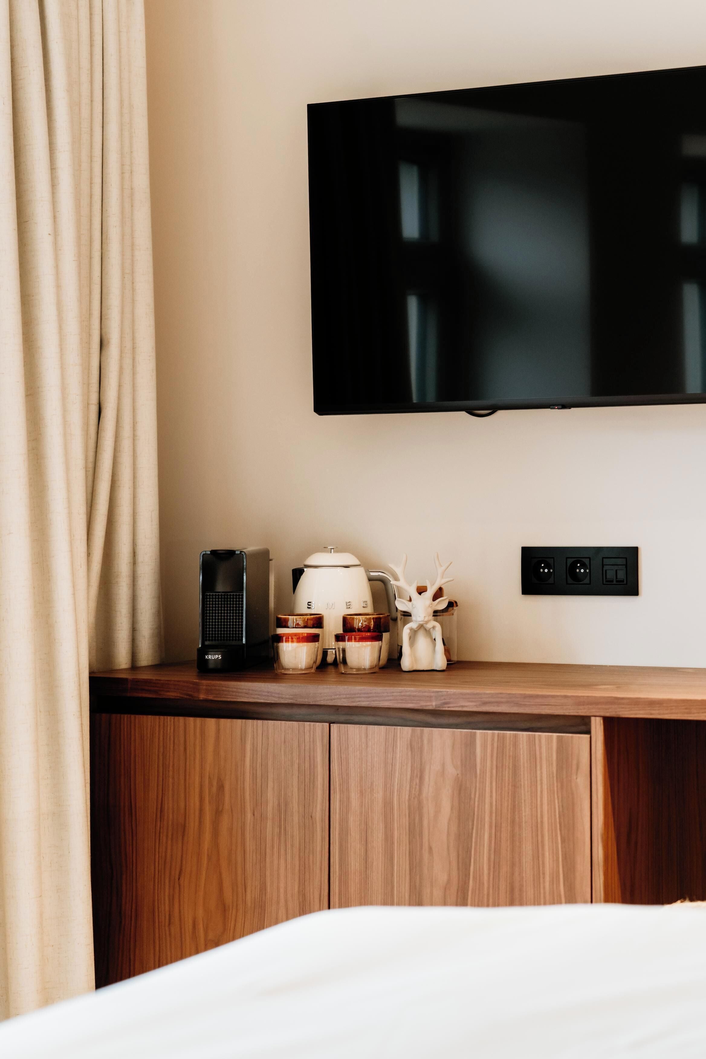 A hotel room with a wooden bedside table, a flat-screen TV mounted on the wall, a coffee maker, and a small decorative item.