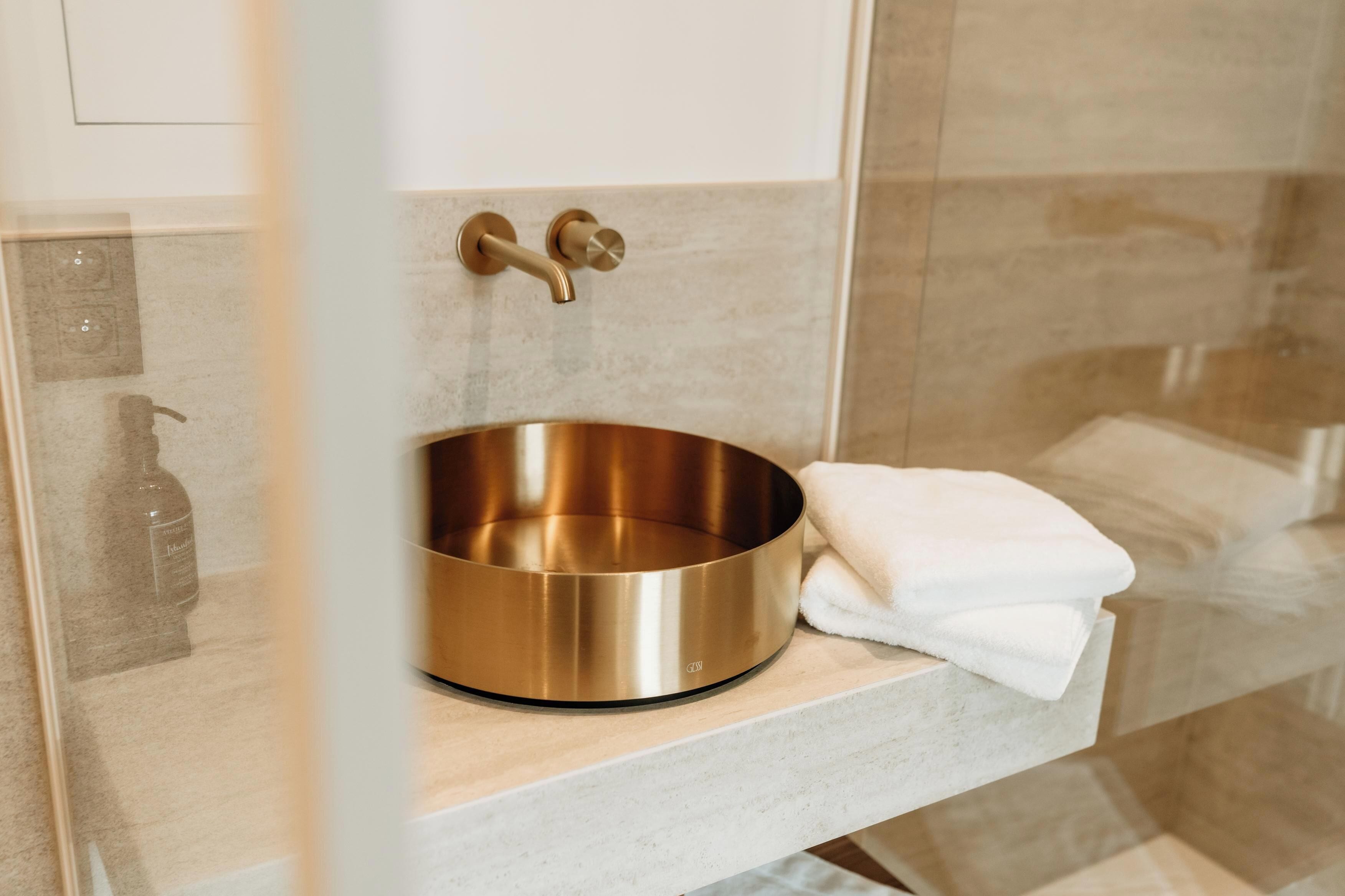 A bathroom with a gold basin on a marble countertop, a towel, and a glass shower door.