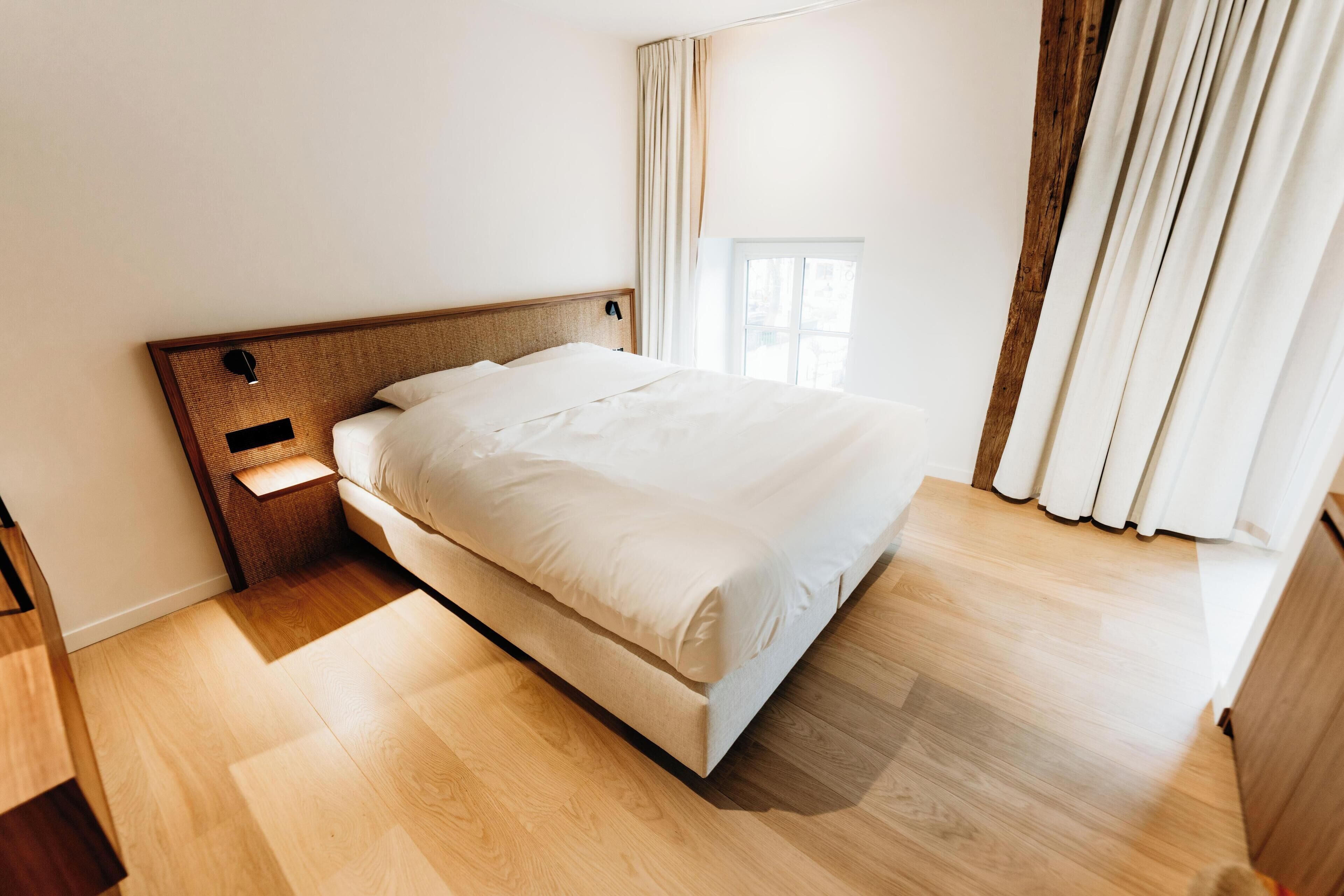 Ein ordentlich bezogenes Bett in einem Zimmer mit Holzdielen, einem Nachttisch und einem Fenster mit Vorhängen.