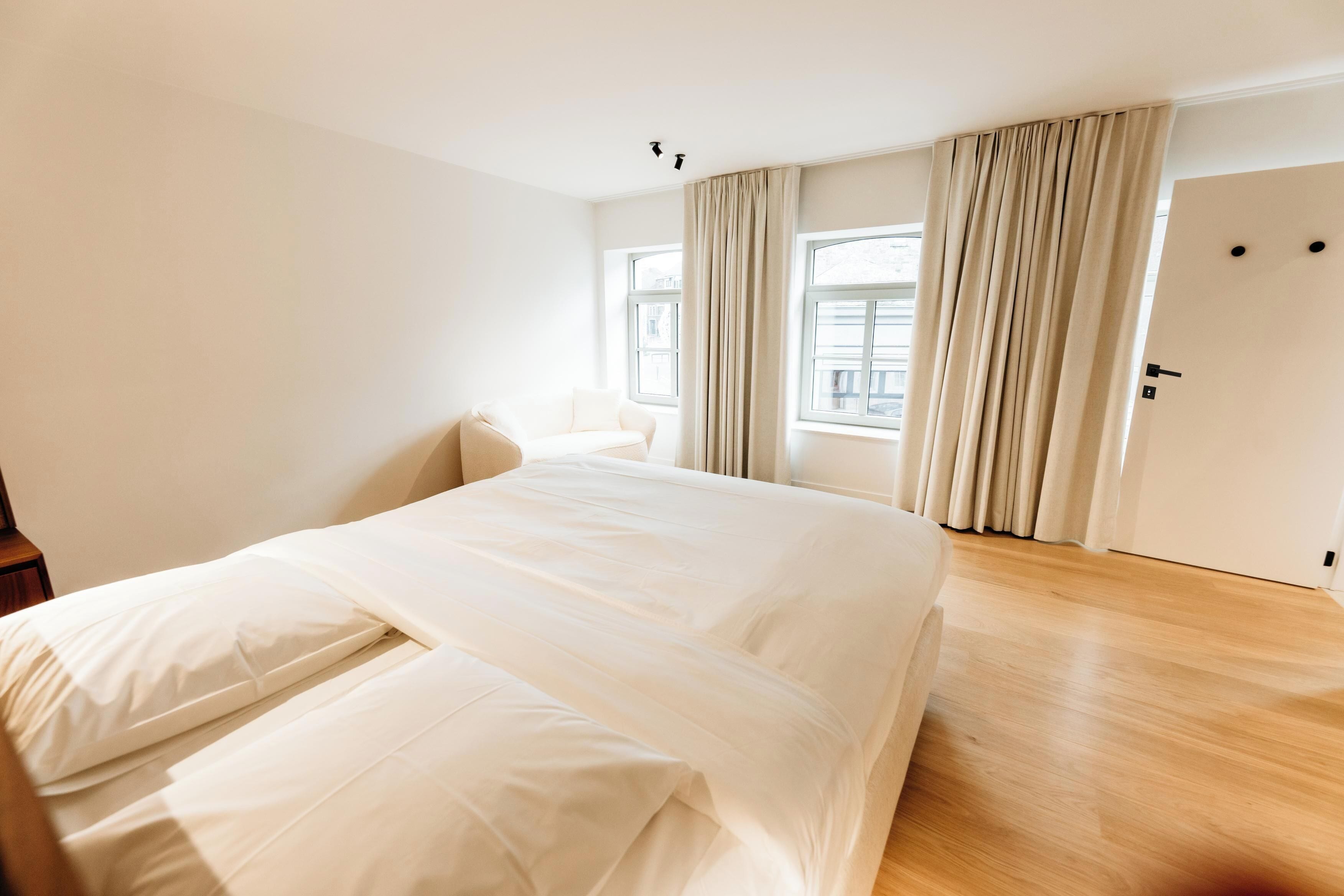 Quarto de hotel com uma cama grande, uma cadeira, piso de madeira e uma porta.