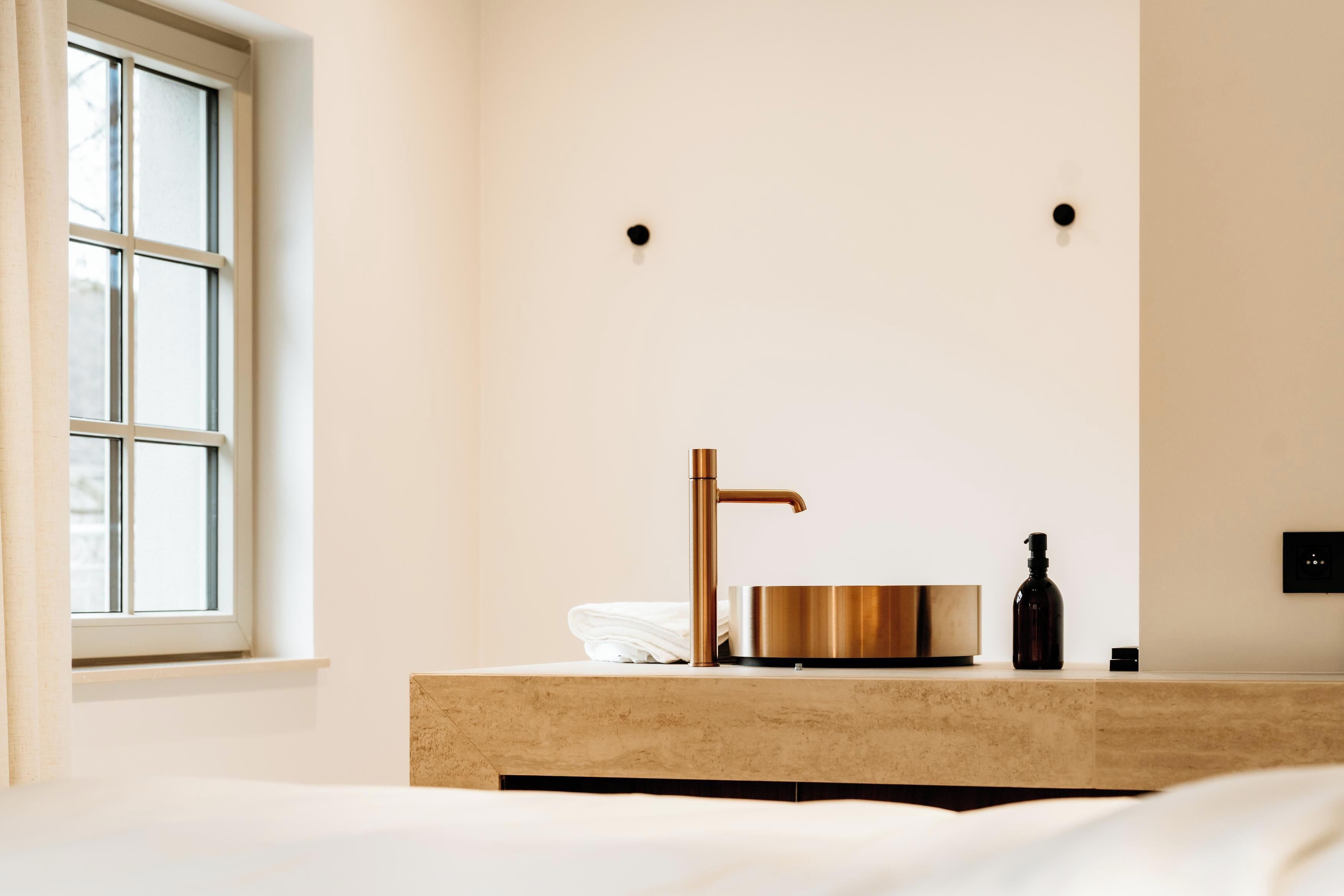 Un bagno moderno con una grande finestra, un rubinetto in rame, un lavabo rotondo e una bottiglia nera sul ripiano.