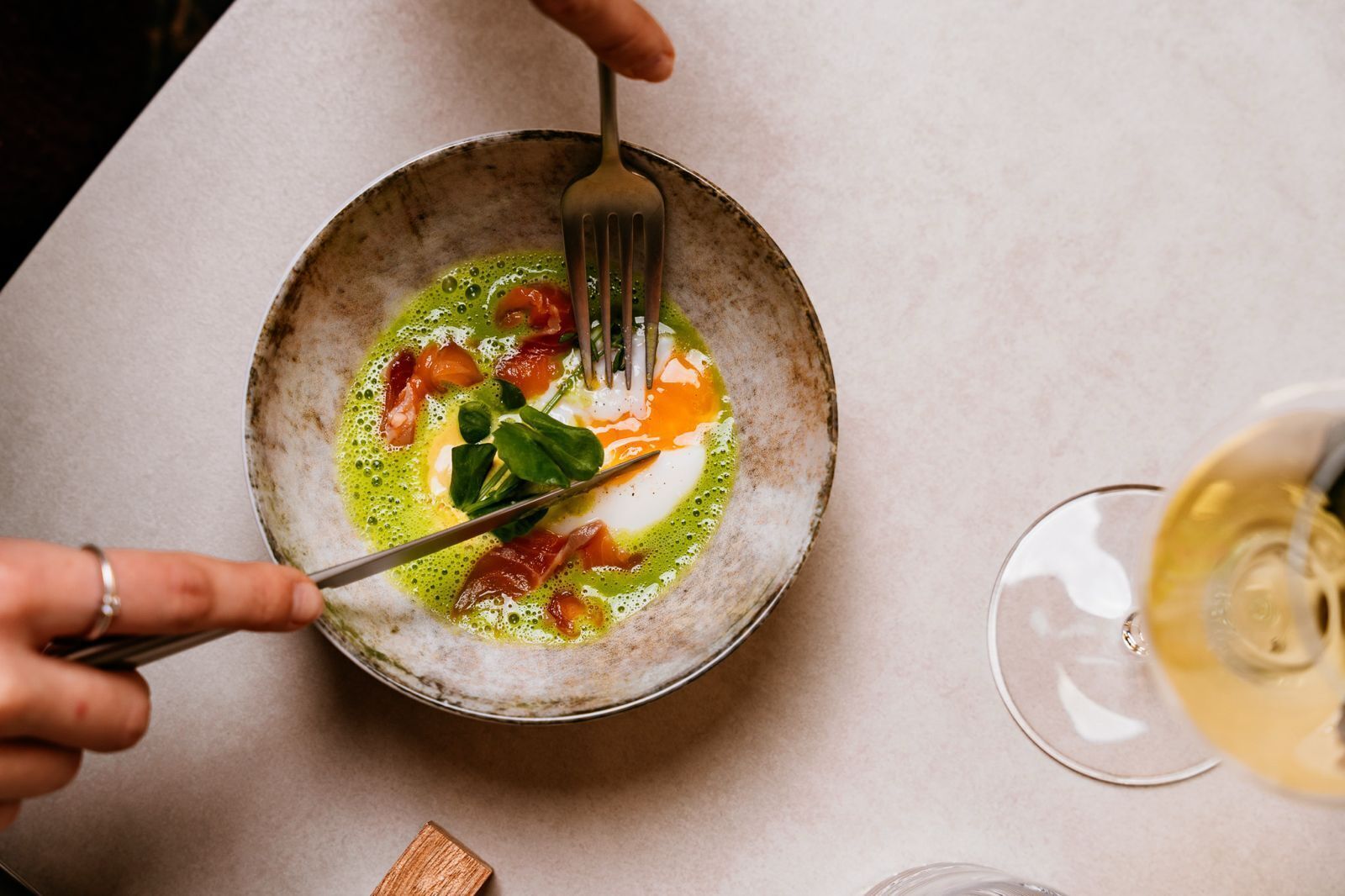 A bowl of food with a fork and knife, a glass of white wine, and a wooden block on a table.