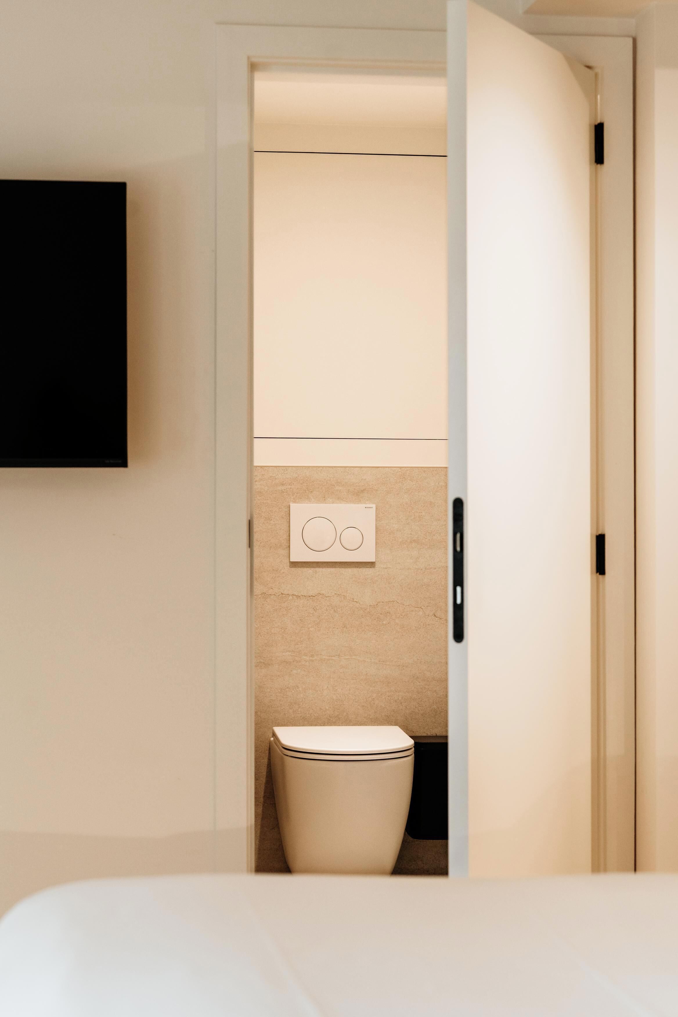 Een hotelkamer met een bed, een aan de muur bevestigde televisie, een toilet en een deur naar een andere kamer.
