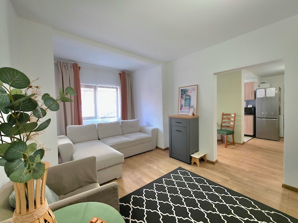 A living room with a sofa, a small table, a plant, and a kitchen area with a refrigerator and microwave.