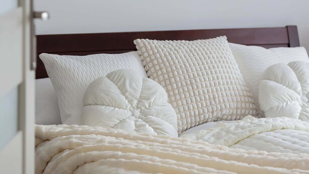 Ein Bett mit weißer Decke und Kissen, ein hölzernes Kopfende und eine Wanddekoration aus Makramee.