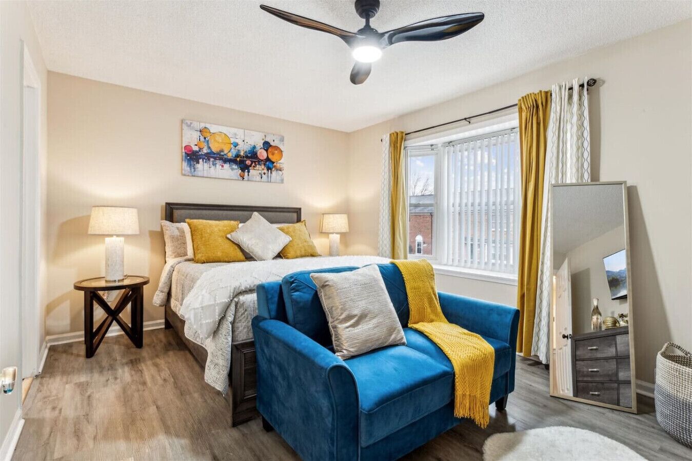 A bedroom with a bed, a blue sofa, a ceiling fan, a window with curtains, and a painting on the wall.