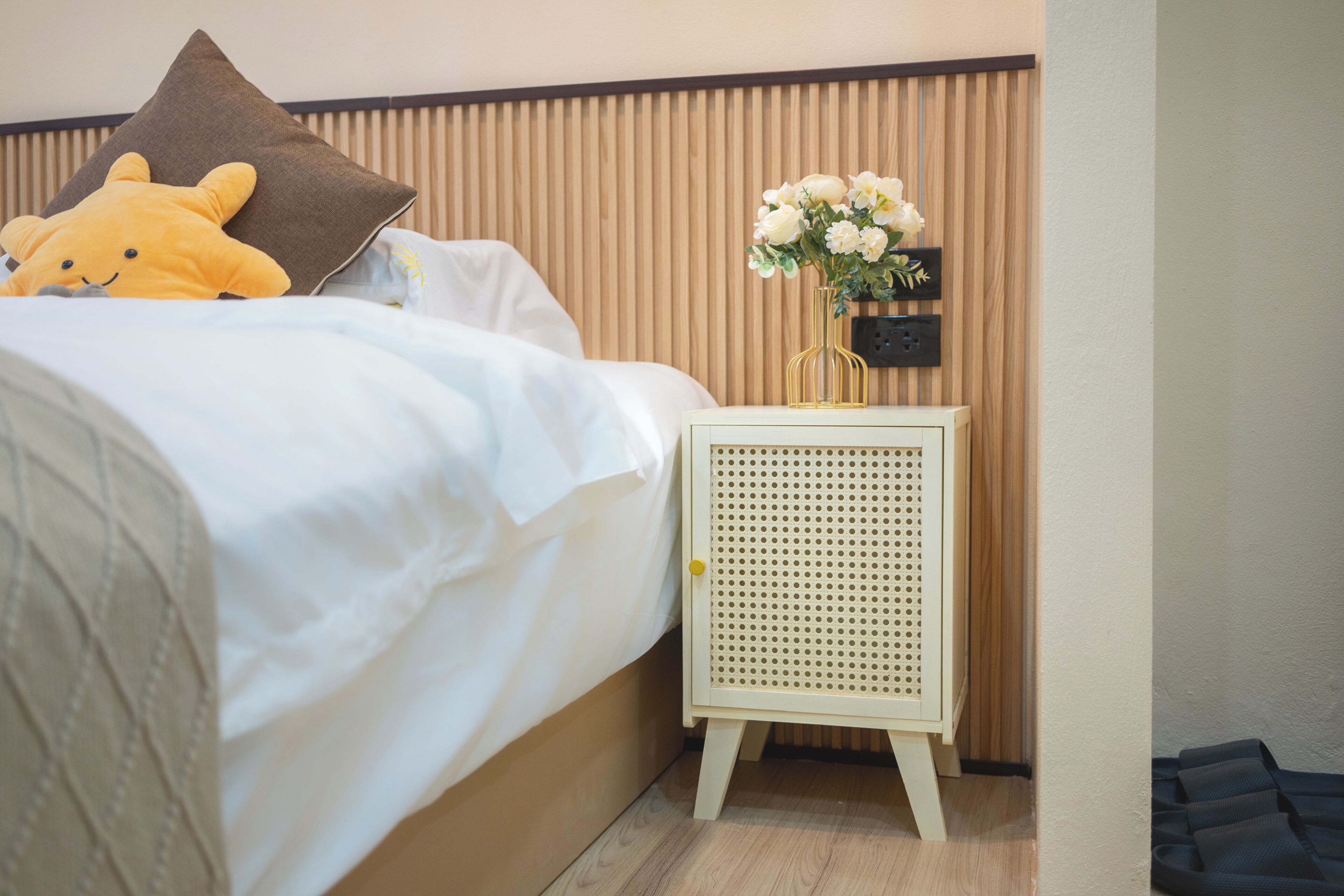 Un lit bien fait, avec un oreiller marron, un jouet en peluche jaune et un vase blanc contenant des fleurs, posé sur une table de chevet.