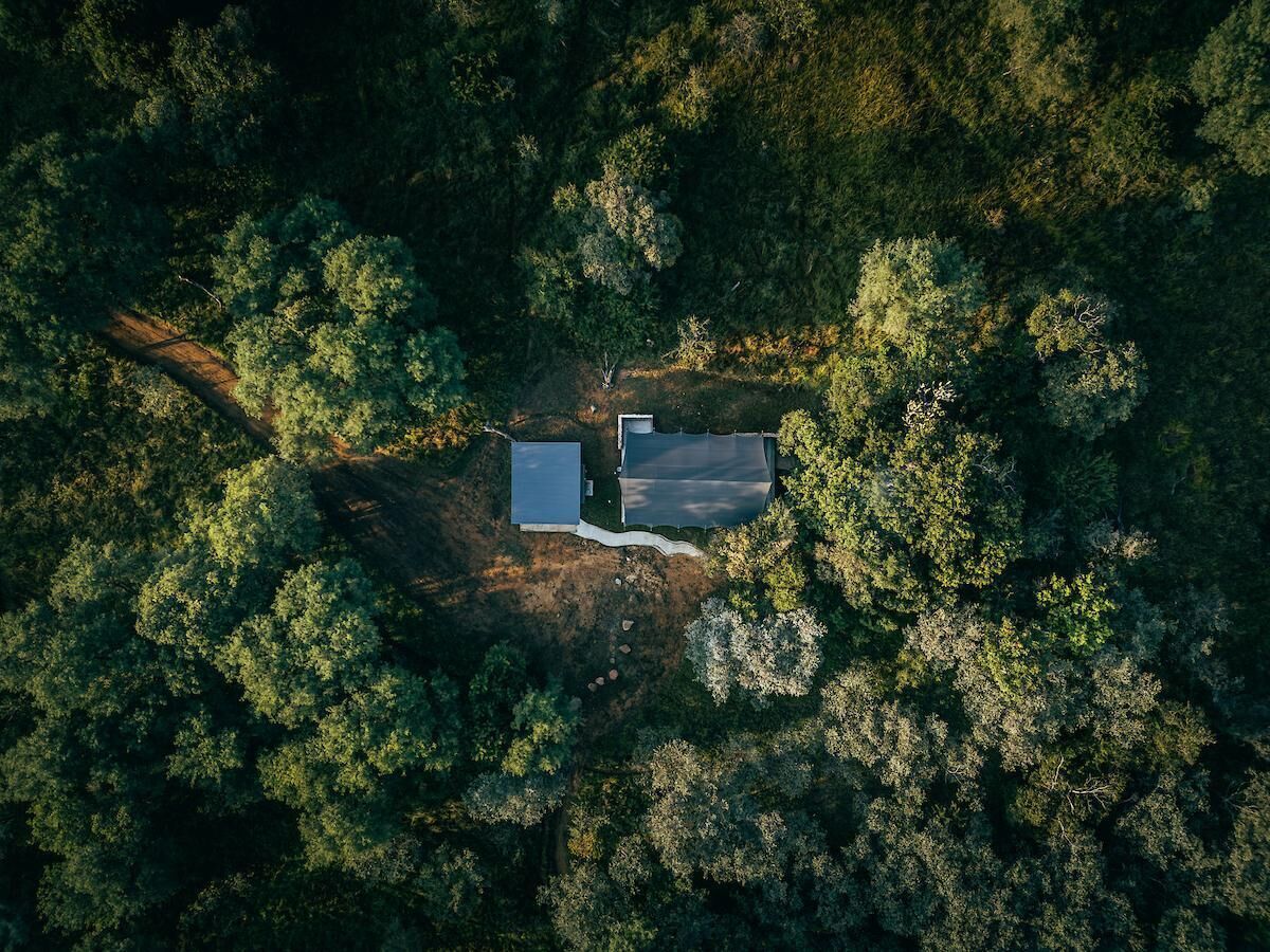 Vista aérea de una propiedad rodeada de bosque denso, con un edificio rectangular de techo plano y una estructura más pequeña adyacente.