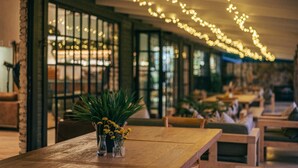 A long wooden table with small glass vases of yellow flowers, set in a dimly lit room with string lights overhead and large windows along one side. - Summerplace Game Reserve (Vaalwater)