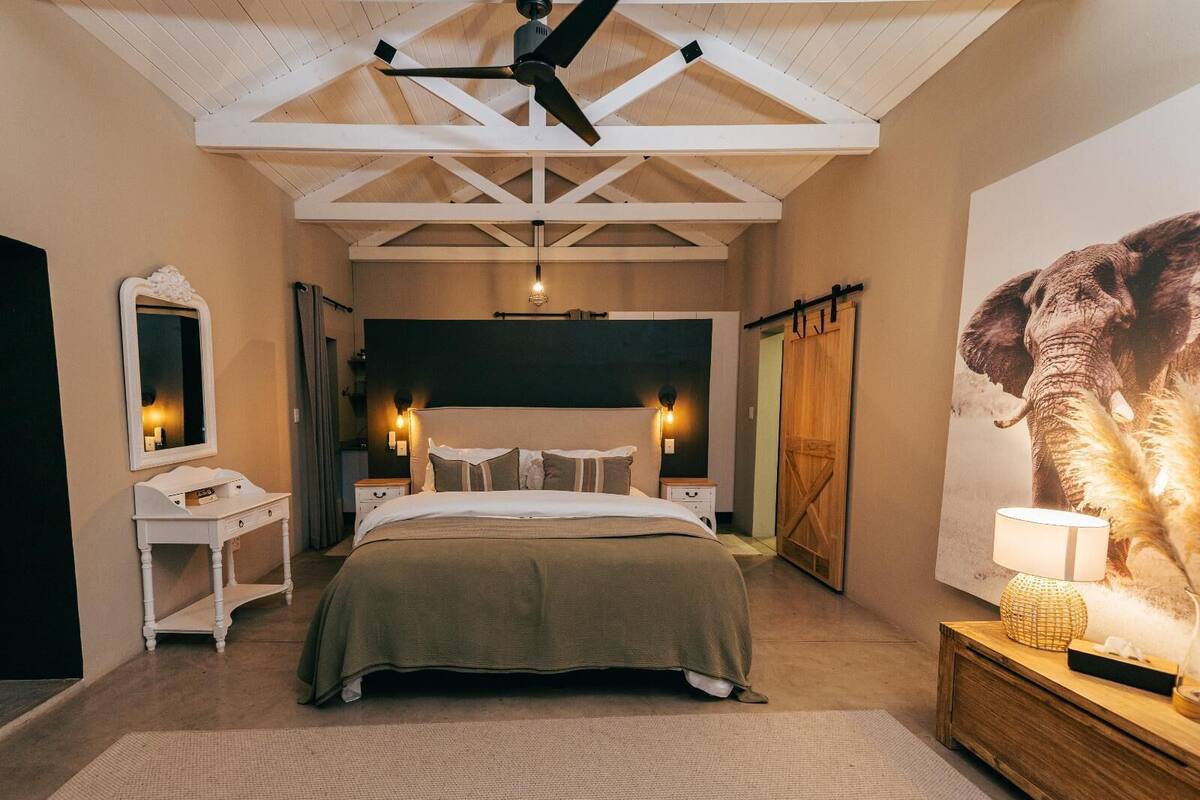 Ein Schlafzimmer mit einem großen Bett, einer Holzzahlende, einem Nachttisch, einer Kommode, einem Spiegel und einem großen Elefantenbild an der Wand.