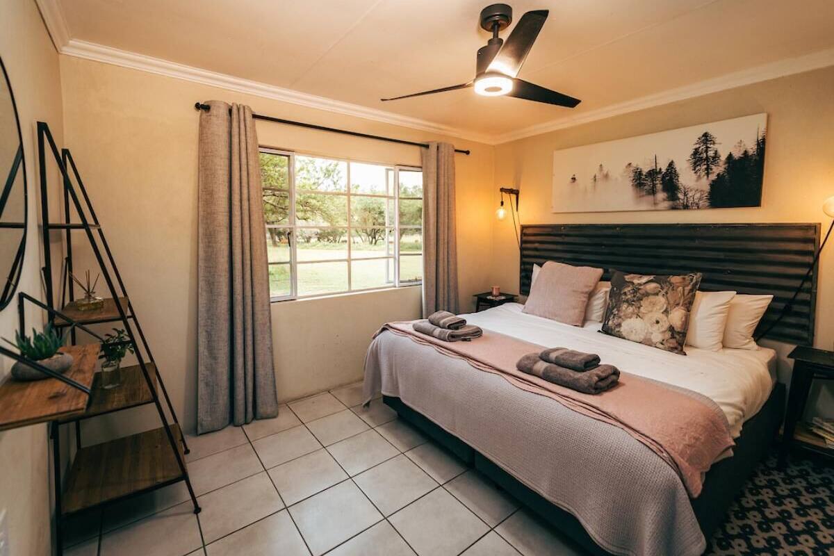 Ein Schlafzimmer mit Bett, Deckenventilator, Fenster mit Vorhängen, Nachttisch und Wandregal mit Dekoration.