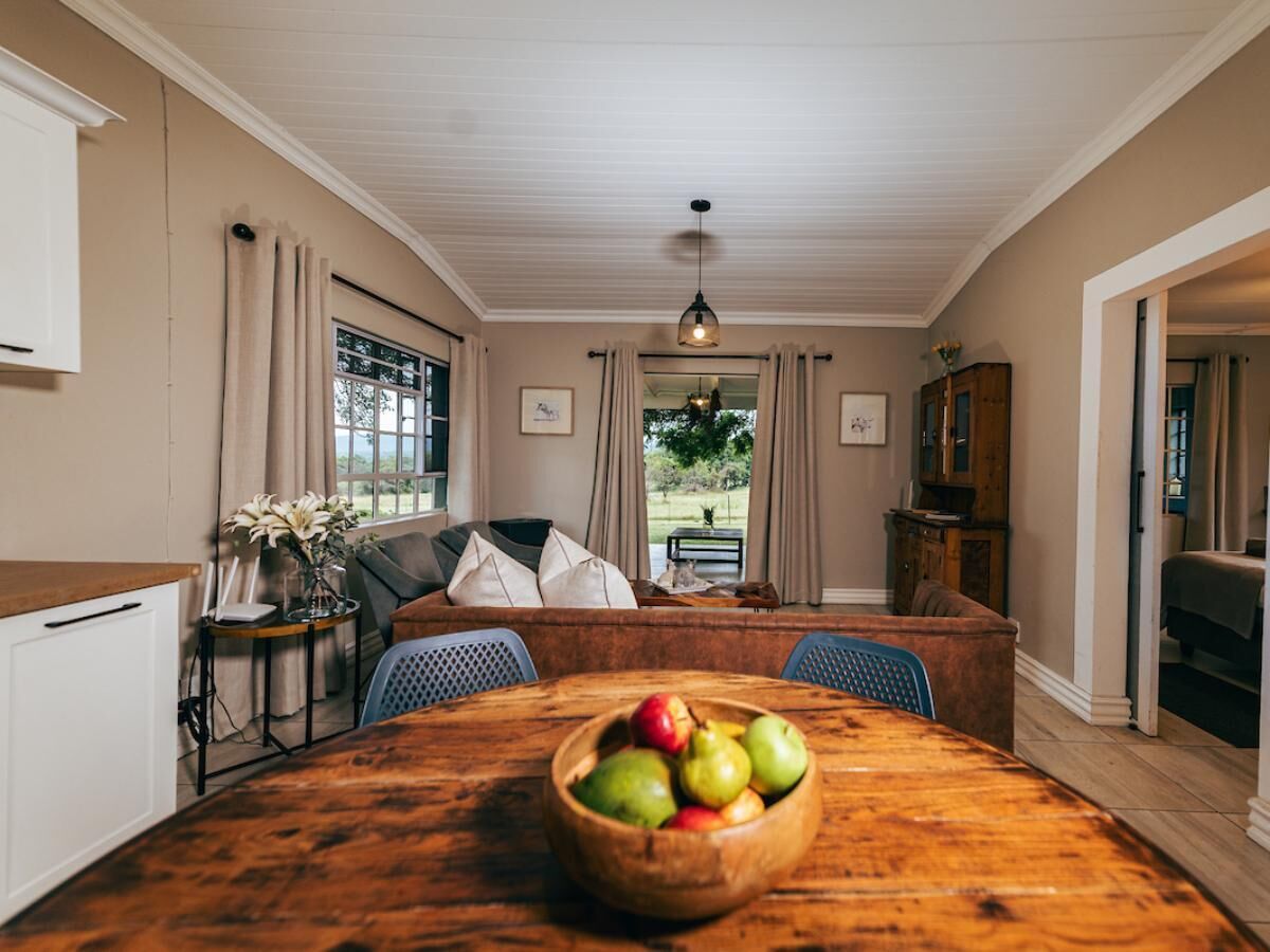 A dining area with a wooden table, a bowl of fruit, and a view of the outdoors through large windows.