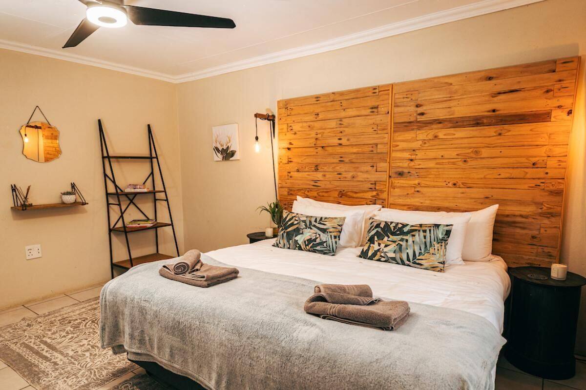 Ein Schlafzimmer mit einem Holz-Kopfteil, einem Bett mit weißen Leinentüchern, einer grauen Tagesdecke und zwei Handtüchern. Gegen die Wand lehnt eine Leiter, ein kleines Regal mit Dekorationsobjekten und eine Wandleuchte.