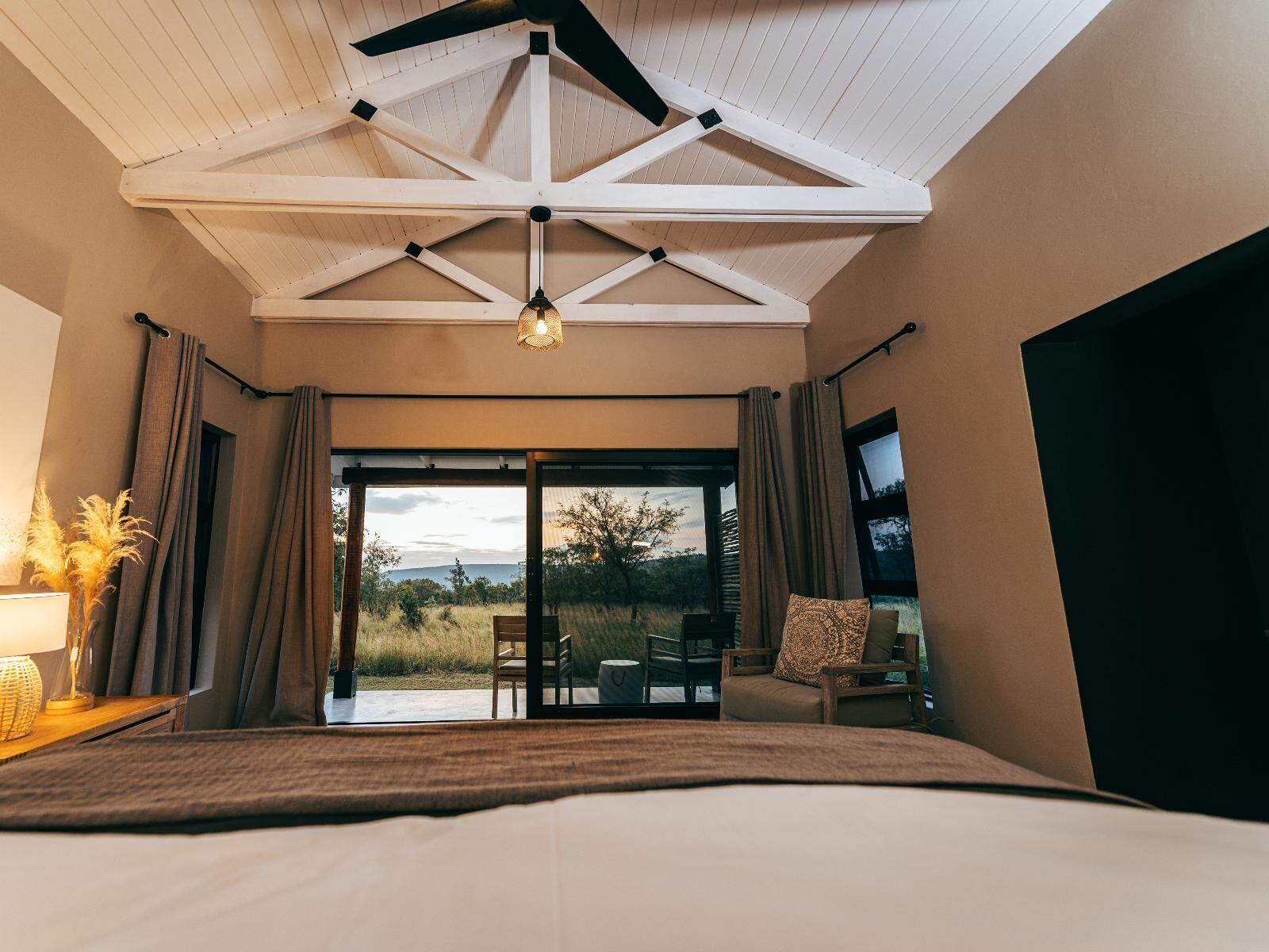 A bedroom with a large bed, a ceiling fan, a view of the outdoors, and a window with curtains.